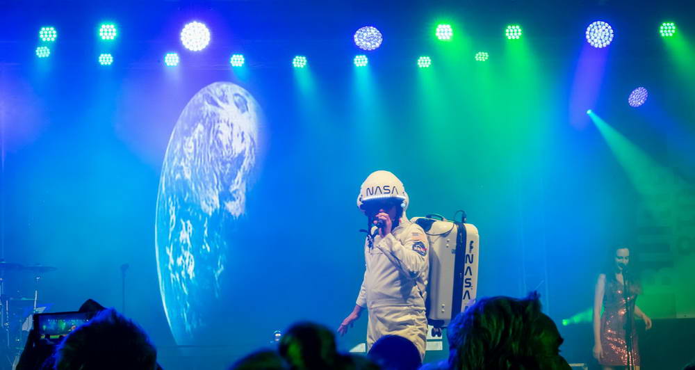 Sänger als Major Tom im Raumfahreranzug. Im Hintergrund auf der Leinwand ist die Erde zu sehen...