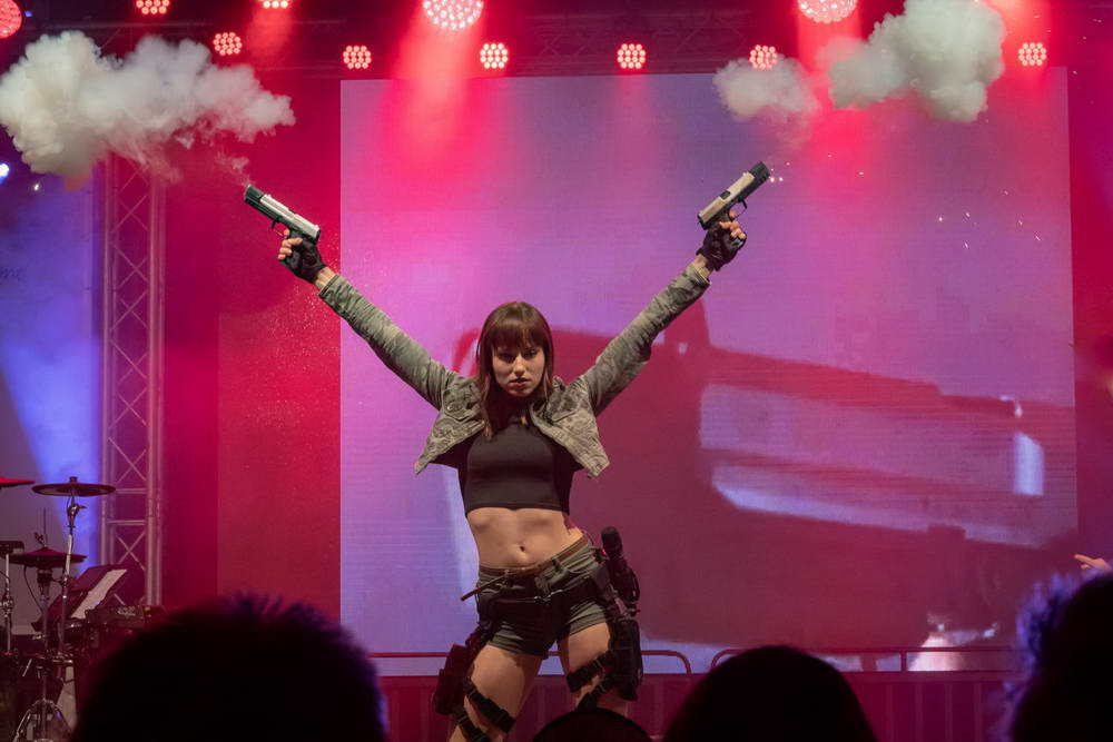 Sängerin im Lara Croft Outfit hält zwei Pistolen in die Luft, aus denen ein Knalleffekt ausgelöst wird. Rauch steigt aus den Pistolen auf.